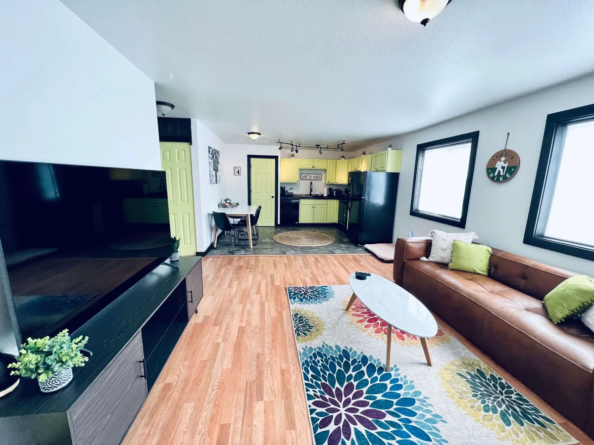 A warm, inviting living area with a brown leather sofa, colorful area rug, green kitchen cabinets, black refrigerator, and a welcoming dining table. The open layout is ideal for immediate relaxation and family gatherings.