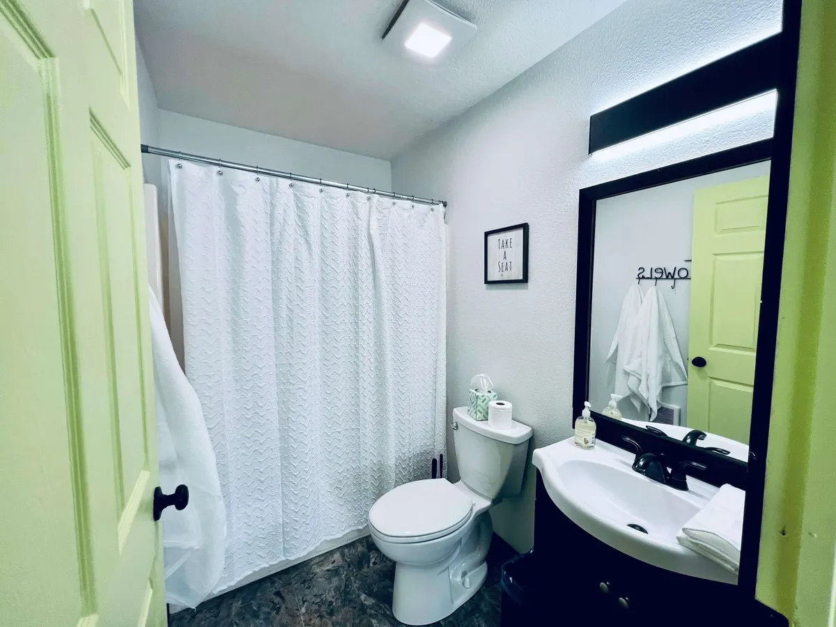 A view of the bathroom includes a toilet, sink, mirror, and shower curtain. The space is fresh and inviting, suggesting both practicality and an opportunity for self-care or a refreshing shower after a long day.
