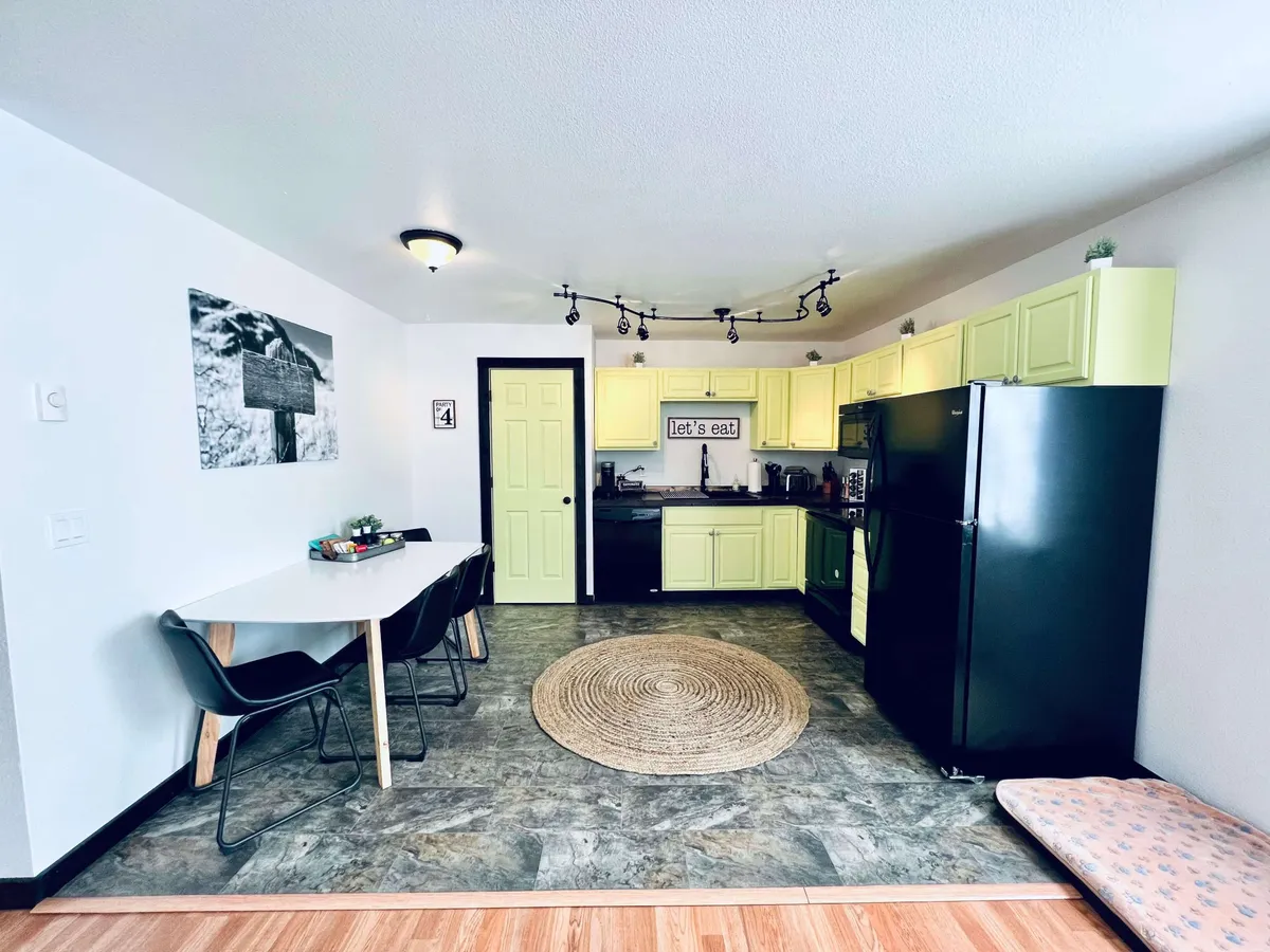 This image displays a bright kitchen with modern cabinetry, a dining table set atop a cozy rug, a sleek black refrigerator, and tasteful wall art. The scene is styled for both home cooking and group meals, with inviting colors and a layout that encourages social interaction.