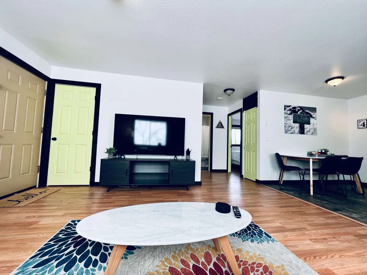 A casual and inviting living space with a television, coffee table, vibrant rug, and striking yellow doors. The area is designed for watching TV, socializing, and enjoying family dinners, offering both comfort and versatility.