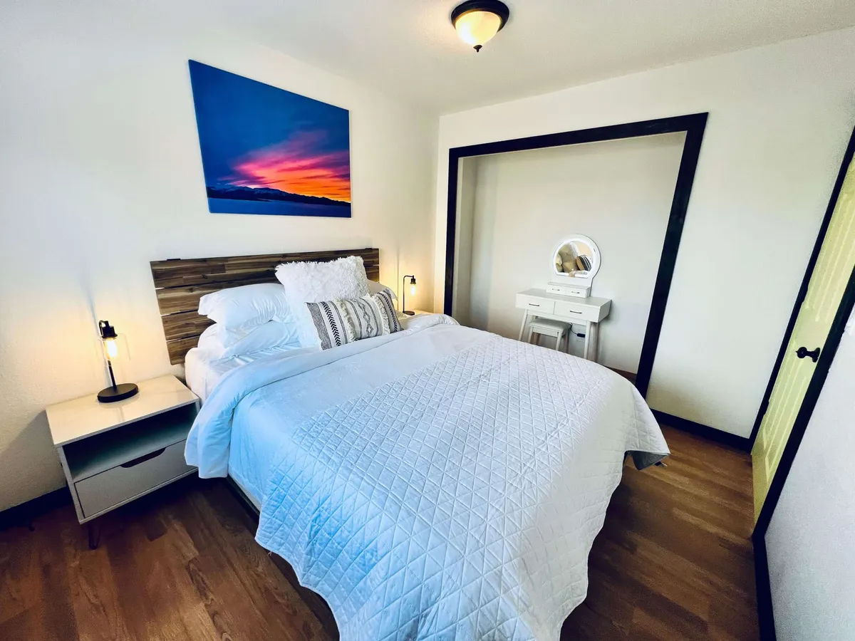 This image depicts a tranquil bedroom featuring a bed with a white quilt, a colorful sunset painting above the headboard, a nightstand with a lamp, and a vanity with mirror. Sunlight filters into the room, creating a peaceful, cozy atmosphere ideal for relaxation or reading.