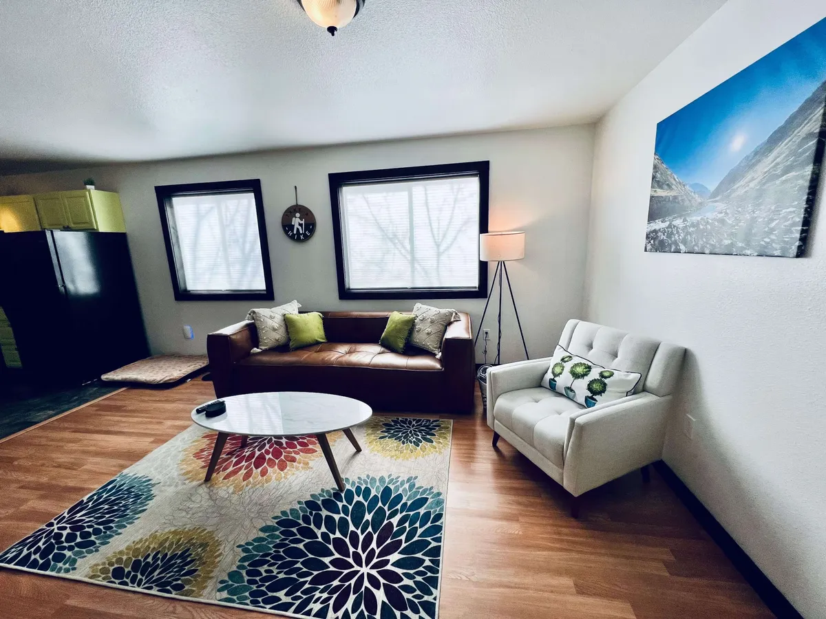 The image showcases a warm, inviting living room with a plush leather sofa, a vibrant area rug, a stylish round coffee table, a cozy floor lamp, and artfully framed mountain landscape artwork on the wall. The space is perfect for relaxation, reading, or socializing, and exudes comfort and charm, making it an ideal gathering spot for guests.