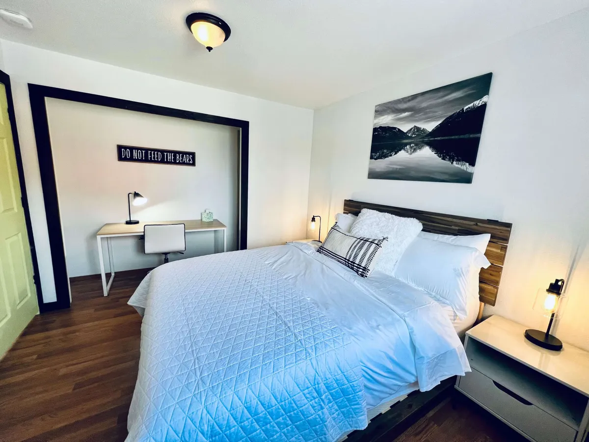 Another bedroom scene shows a comfortable bed with white bedding, a framed mountain landscape photograph above the headboard, and a desk with a lamp and chair. The room is designed for relaxation as well as quiet activities like reading or working, offering a versatile and restful environment.